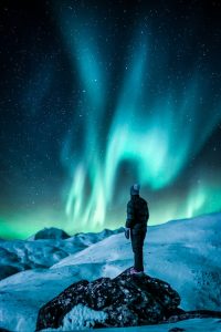 A lone figure stands on a snowy slope, gazing at the vibrant Northern Lights under a starry sky.